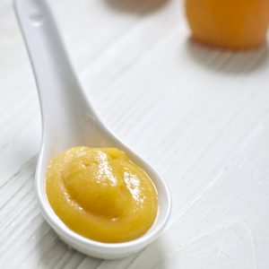 白汤匙充满了浓密的杏仁和梨婴儿食品泥。汤匙坐在白色木制板上，背景上有一些新鲜的杏子。