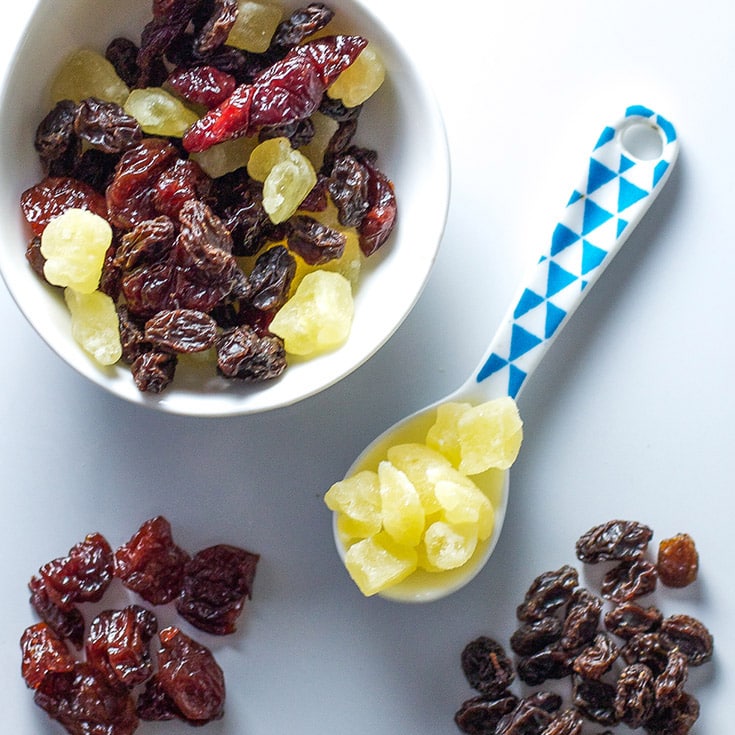 A bowl of dried fruit mix with a toddler spoon on a white background.