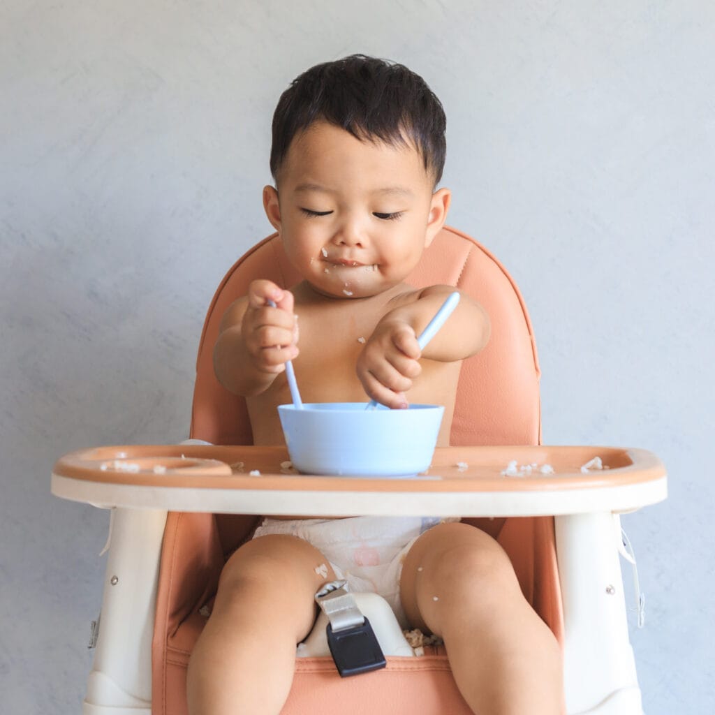 小宝宝坐在高高的椅子上，用两个汤匙吃。