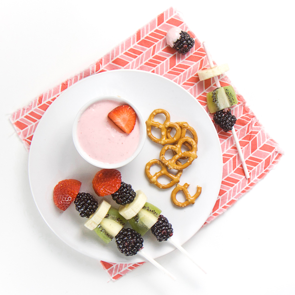在桃红色餐巾的圆的白色板材，小的小孩在板材的碗充满草莓乳脂干酪蘸，果子kababs和一把小少数椒盐脆饼