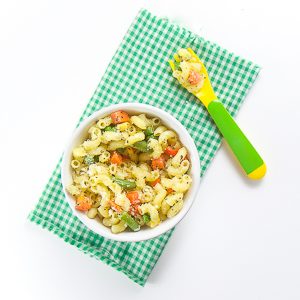 small white bowl filled with pasta and vegetables. Sits on top of a green and white checkered napkin with a green and yellow fork filled with pasta