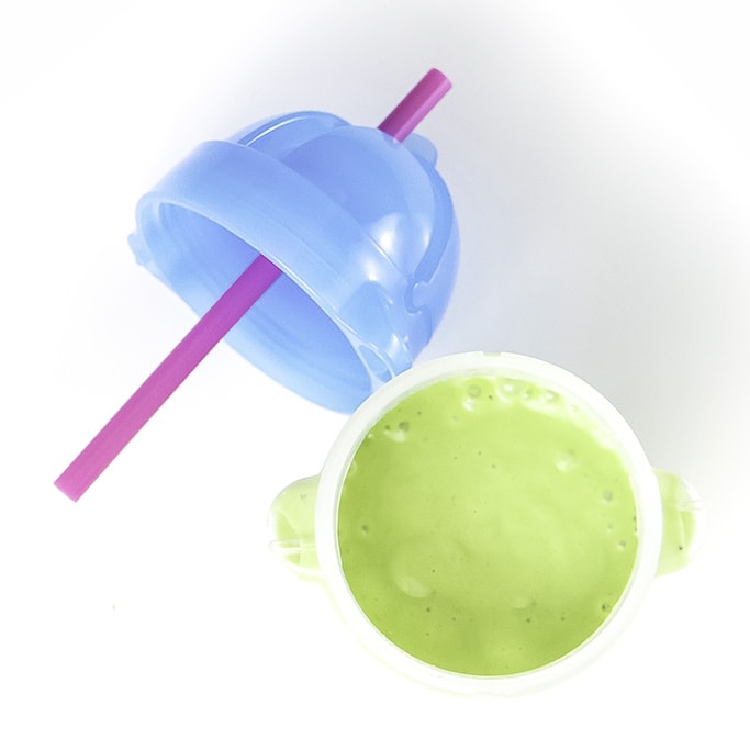 透明的吸管杯，装满了绿色的鳄梨冰沙，可为婴儿和幼儿坐在白色的表面上。