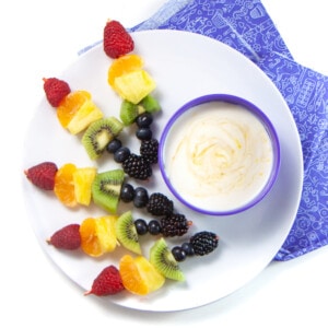 一碗白色盘子，上面有几个彩虹色的水果串，一碗酸奶蘸酱，紫色的儿童餐巾在白色背景上。