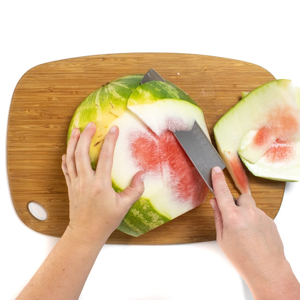 一个木制切菜板，侧面有一条西瓜，一只手将西瓜的果皮修剪起来。