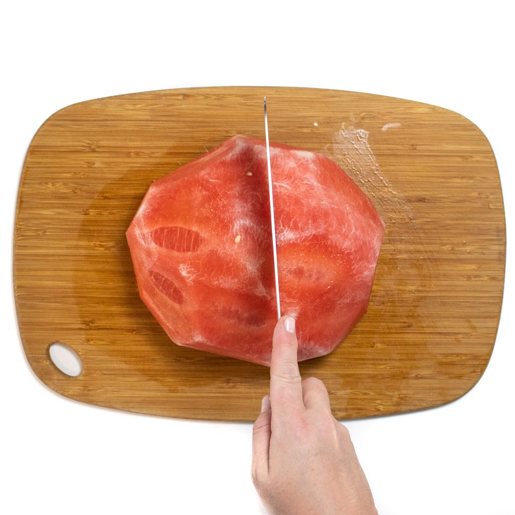 一半的剥皮西瓜在木制切菜板上，手握着一把刀将其切成两半。