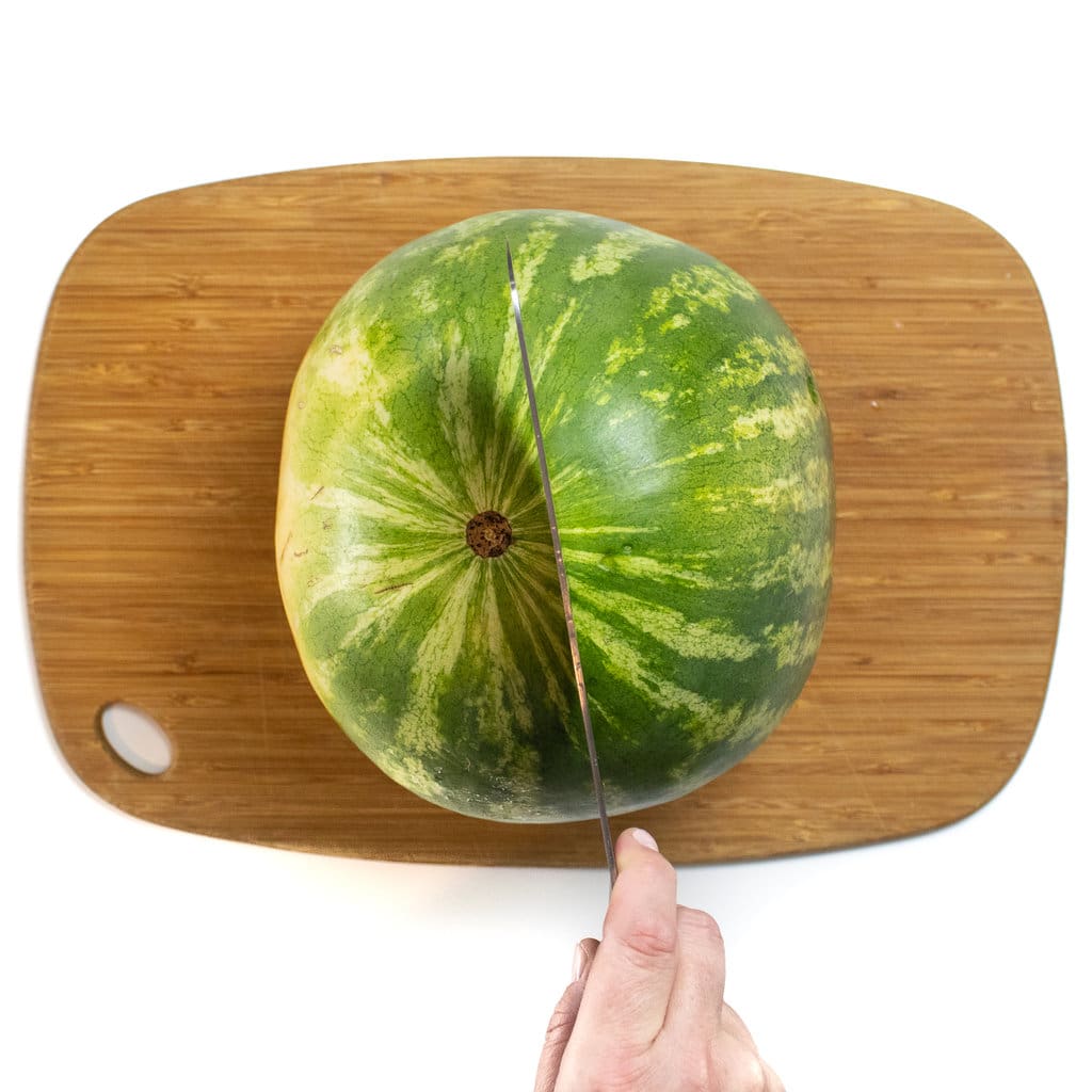 一个西瓜坐在切菜板上，用刀将其切成两半。