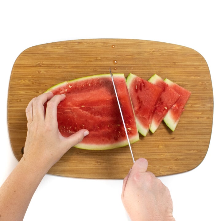 用两只手拿着一只手拿着刀和切片西瓜在木制切菜板上的西瓜楔子。