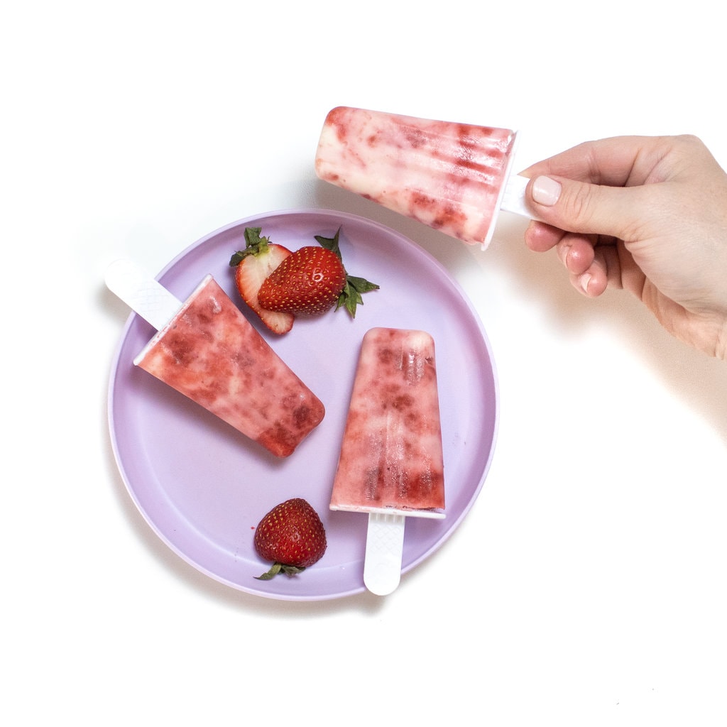 一个孩子紫色的盘子，上面有两个草莓酸奶冰棍和切成薄片的草莓，一只手拿着另一只草莓酸奶冰棒在白色背景上。