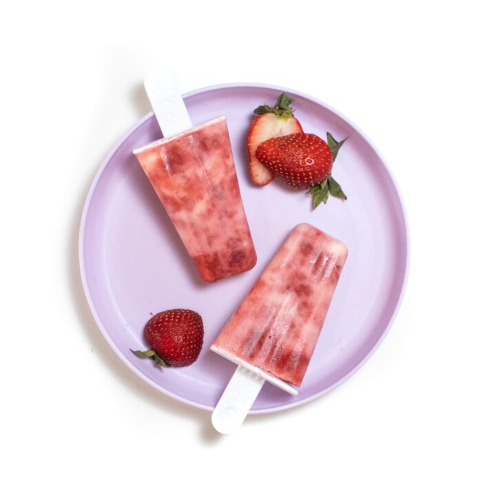 一个紫色的孩子盘，上面有两个草莓酸奶冰棒和在白色背景上切成薄片的草莓。