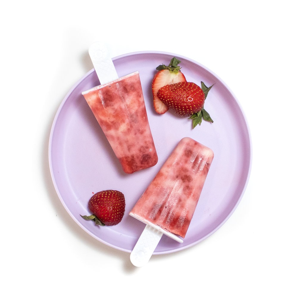 一个紫色的孩子盘，上面有两个草莓酸奶冰棍，并在白色背景上切成薄片的草莓。