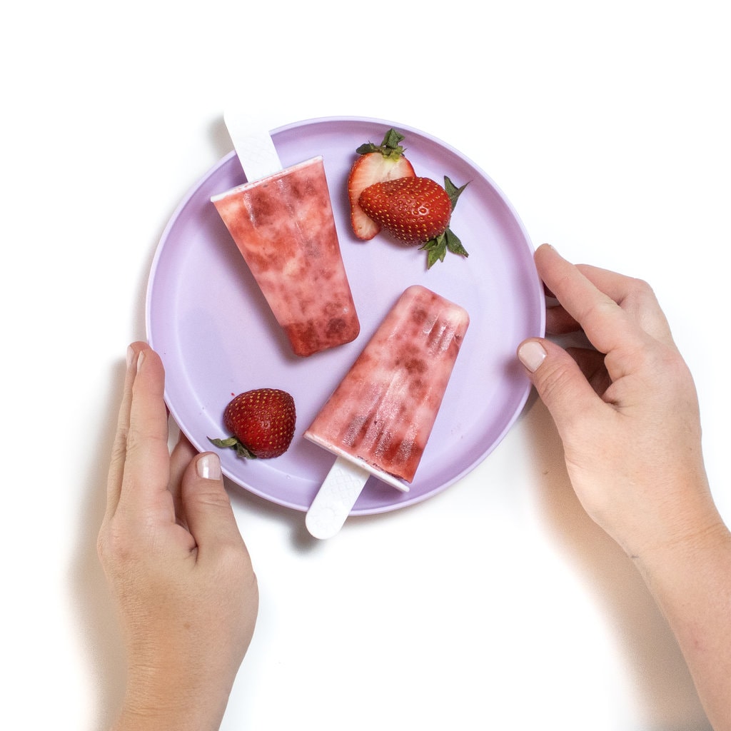 紫色的儿童盘，两只手将其握在草莓酸奶冰棍和切成薄片的草莓中。
