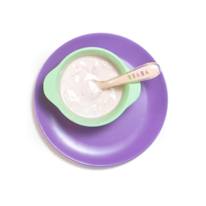 一个桃子婴儿勺子放在蓝绿色的婴儿碗上，上面放着矮胖的西瓜和酸奶酱，坐在紫色的盘子上，靠在紫色的盘子上。