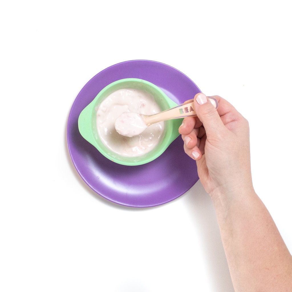 一只手拿着一个小勺子，上面放着矮胖的西瓜泥和酸奶，在蓝绿色的婴儿碗和紫色的婴儿板上。