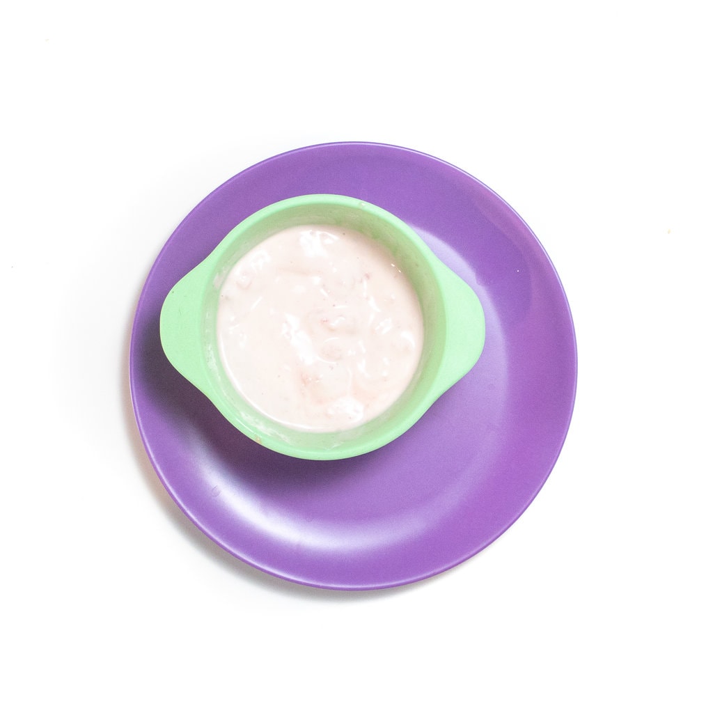 蓝绿色的婴儿碗，上面放着矮胖的西瓜和酸奶泥，在紫色的盘子上靠在白色背景上。