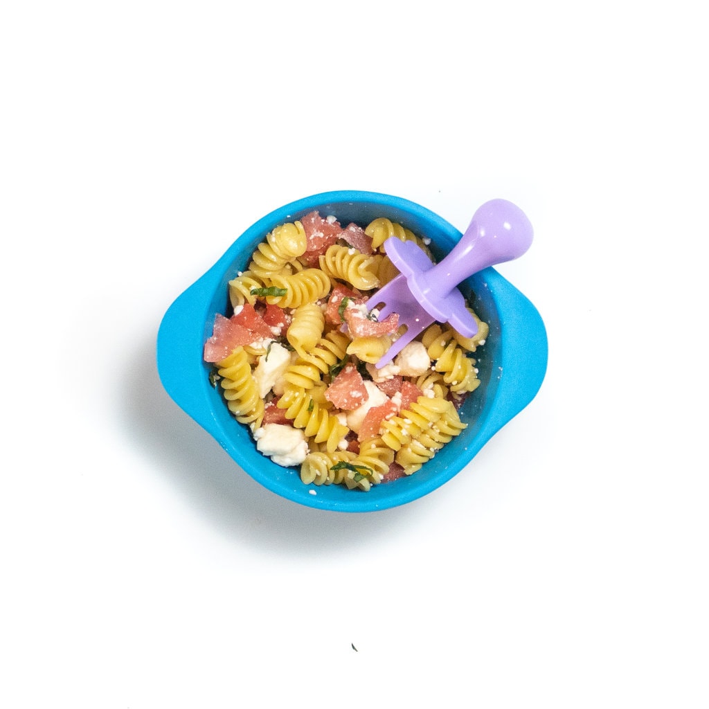 一个蓝色的婴儿碗在白色背景上，碗充满了面食，西瓜，羊乳酪和罗勒，里面放着紫色的婴儿叉。
