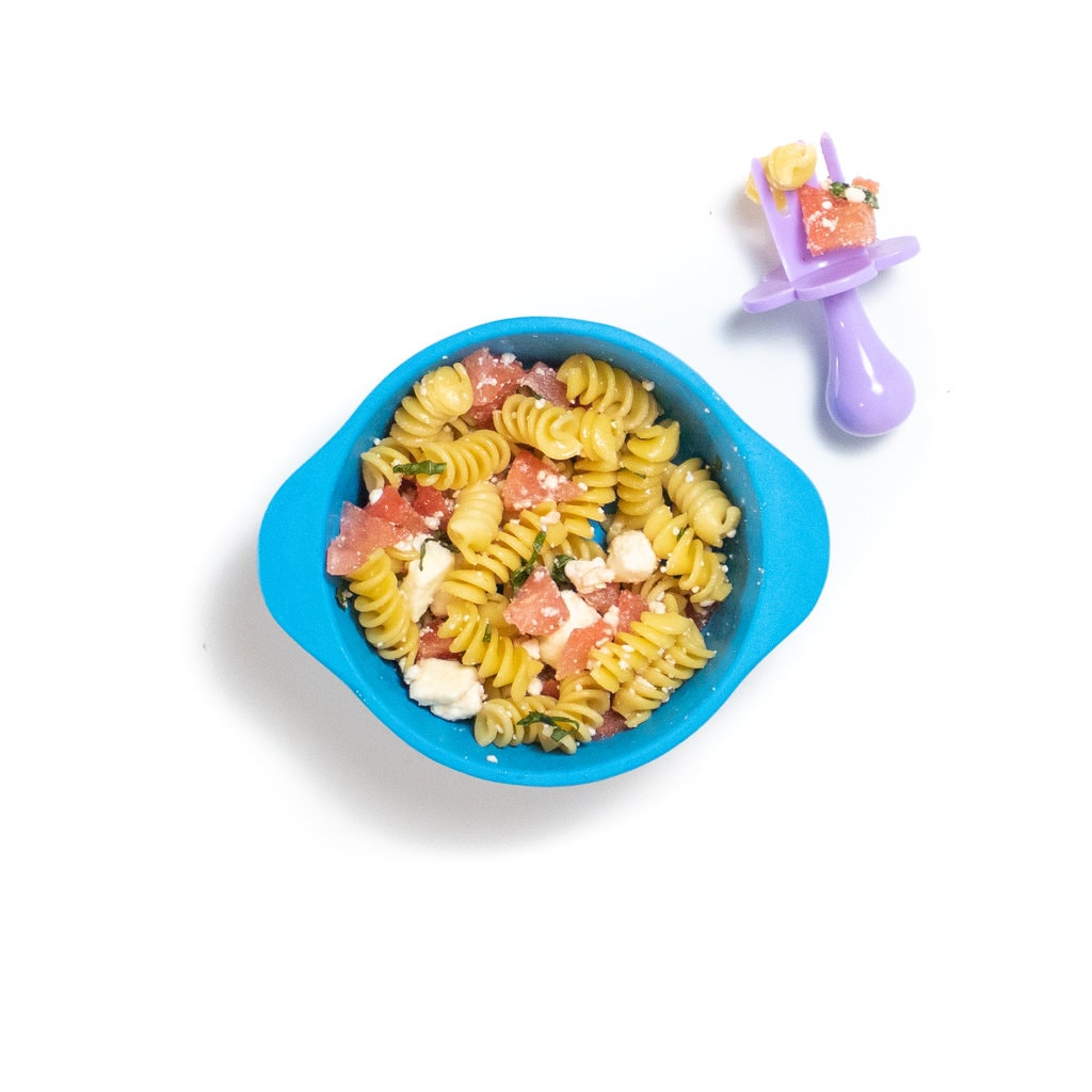 一个蓝色的婴儿碗，里面充满了意大利面，西瓜，羊乳酪和罗勒在白色背景上，紫色的婴儿叉坐在侧面。