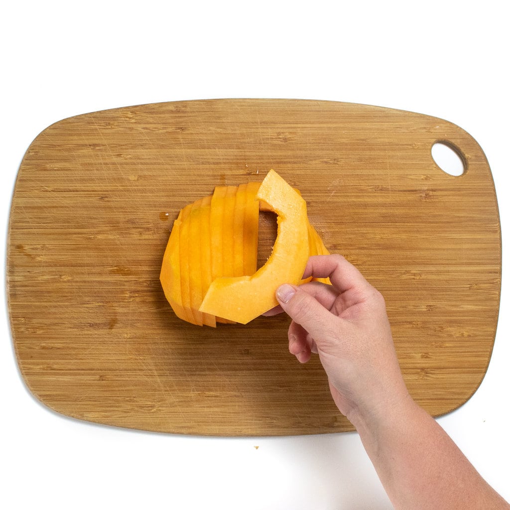 一只手在木制切菜板上方拿着一块薄薄的哈密瓜在白色背景上。