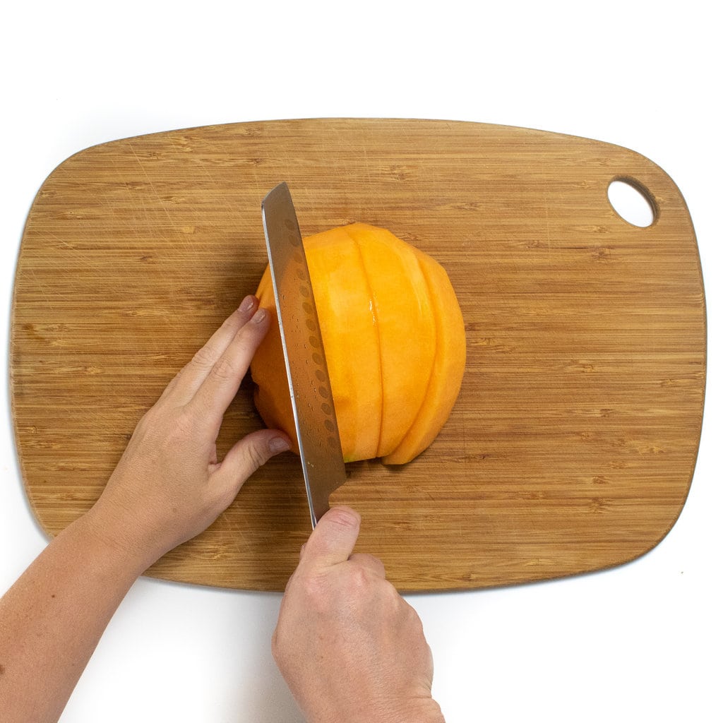 双手握着哈密瓜将其切入木制切菜板上的厚楔形，在白色的背景下。
