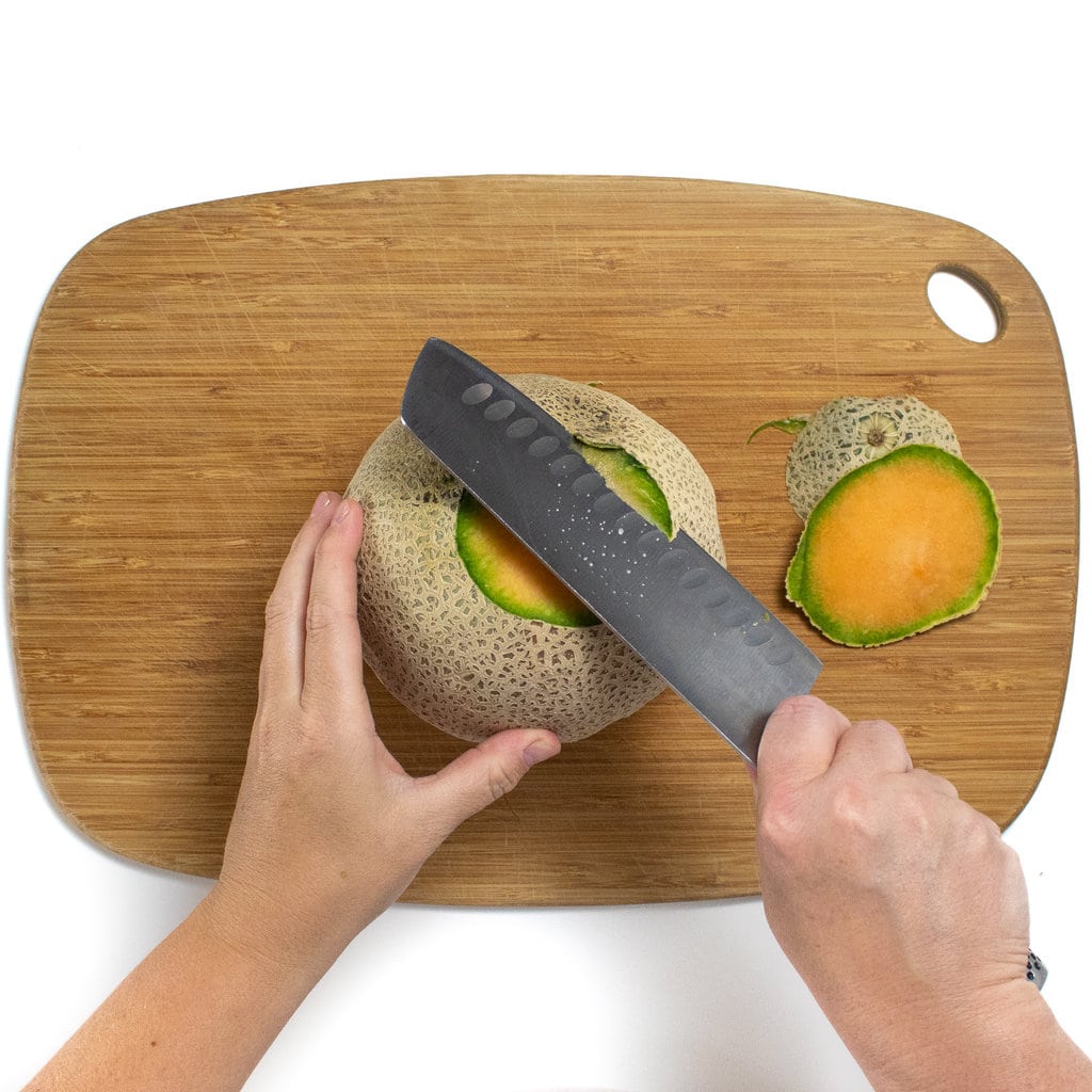 两只手用刀握住哈密瓜，切断了切菜板上哈密瓜肉周围的皮肤。