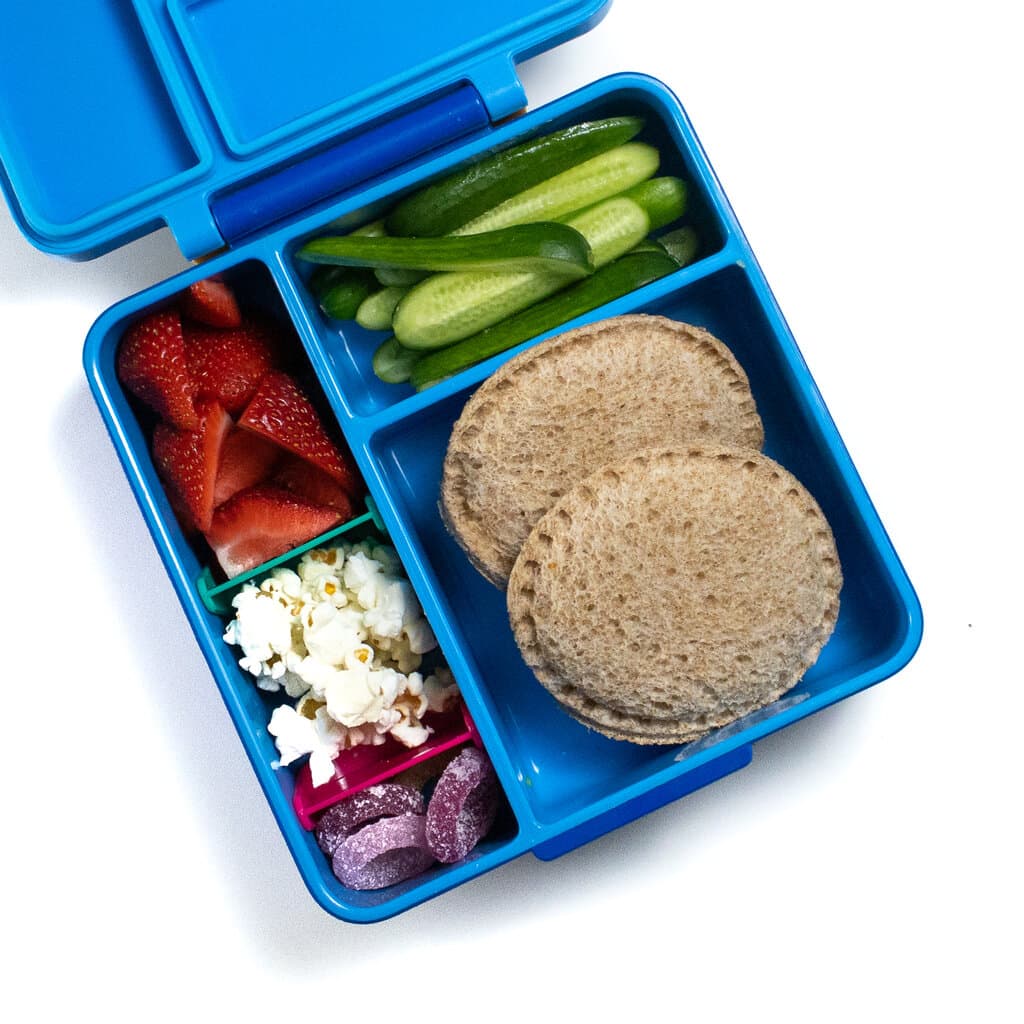 一个蓝色午餐盒，里面装满了两个不可分割的三明治，黄瓜，草莓，爆米花和胶状小吃。