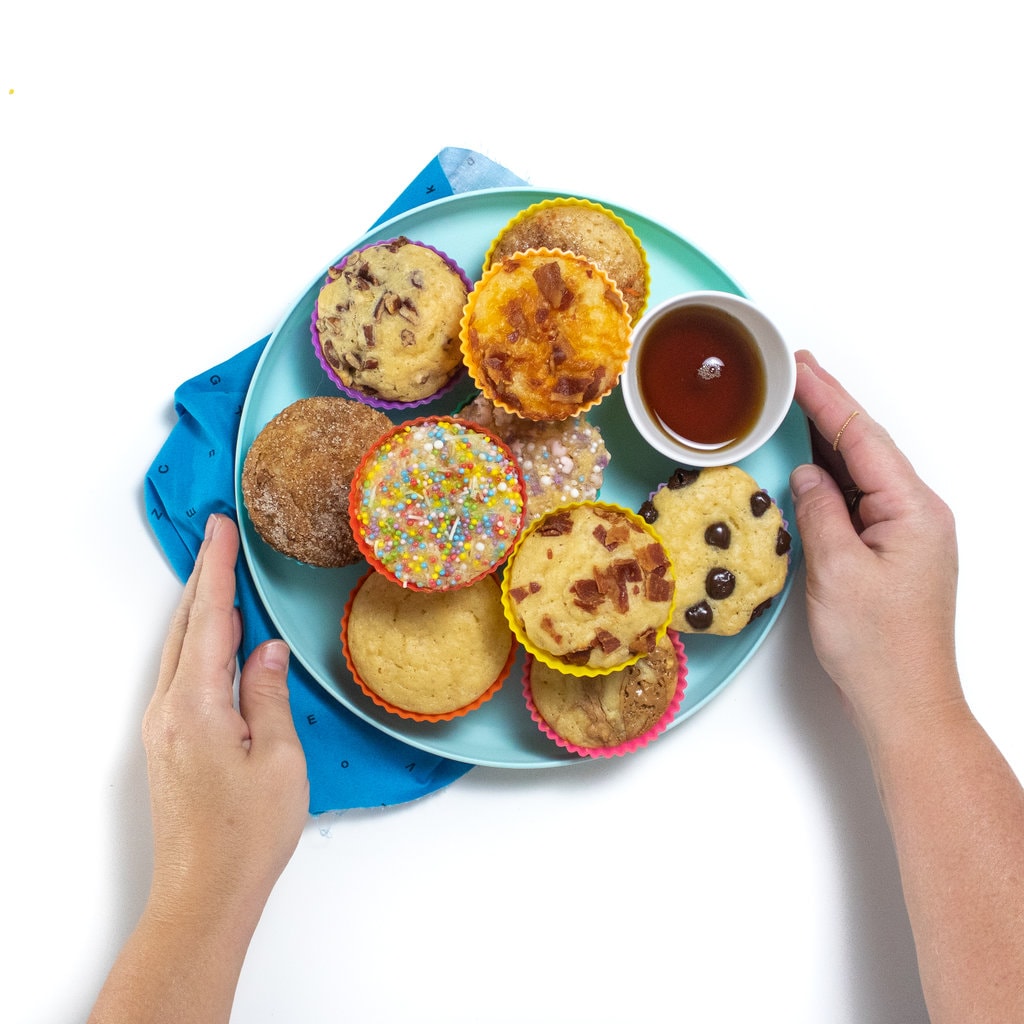 Two hands holding a blue plate full of pancake muffins with different toppings and a small bowl of maple syrup.