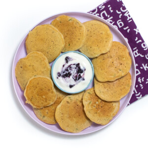 一个装满西葫芦煎饼的紫色儿童盘，里面有一小碗酸奶和蓝莓酱，我可以看到带有紫色餐巾纸的白色背景。