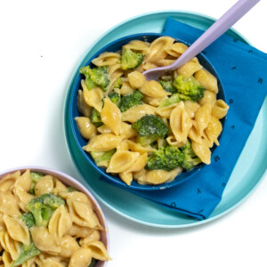 一个蓝色的儿童盘子和碗，上面有蓝色的餐巾纸和紫色叉，碗里有西兰花Mac和奶酪。旁边是一个粉红色的碗，里面装满了西兰花Mac和奶酪。
