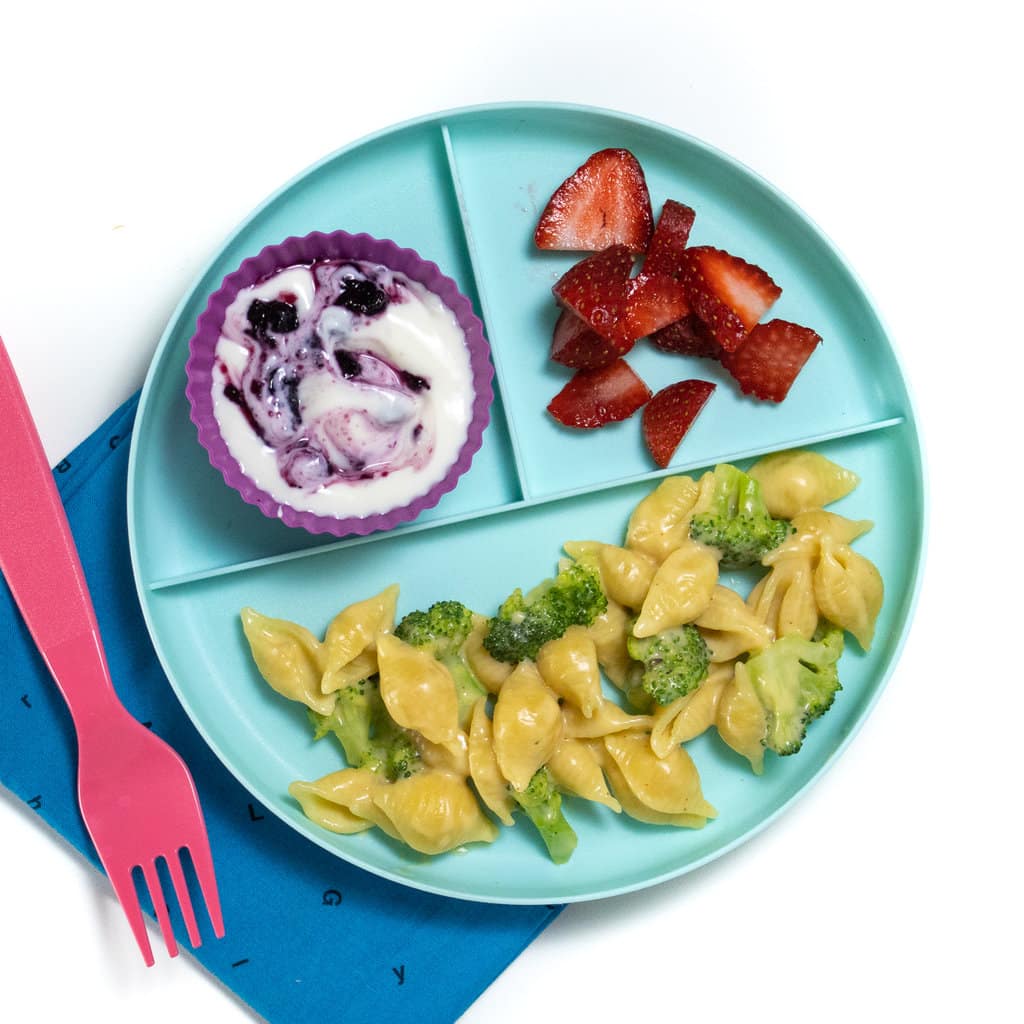 孩子们的三片盘子配西兰花麦克斯和奶酪，切成薄片的草莓和酸奶在白色背景上，带有蓝色的餐巾纸和粉红色的叉子。