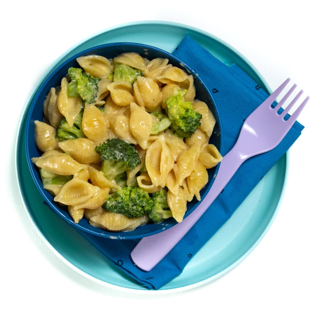 一个蓝色盘子和一个装满西兰花的Mac＆奶酪的蓝色碗，上面有紫色叉，上面放着Mac＆Cheese在白色背景上，带有蓝色的餐巾纸。