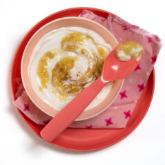 一个粉红色的孩子碗和盘子，上面有粉红色的餐巾纸，里面装满了香蕉酸奶，上面放着勺子。