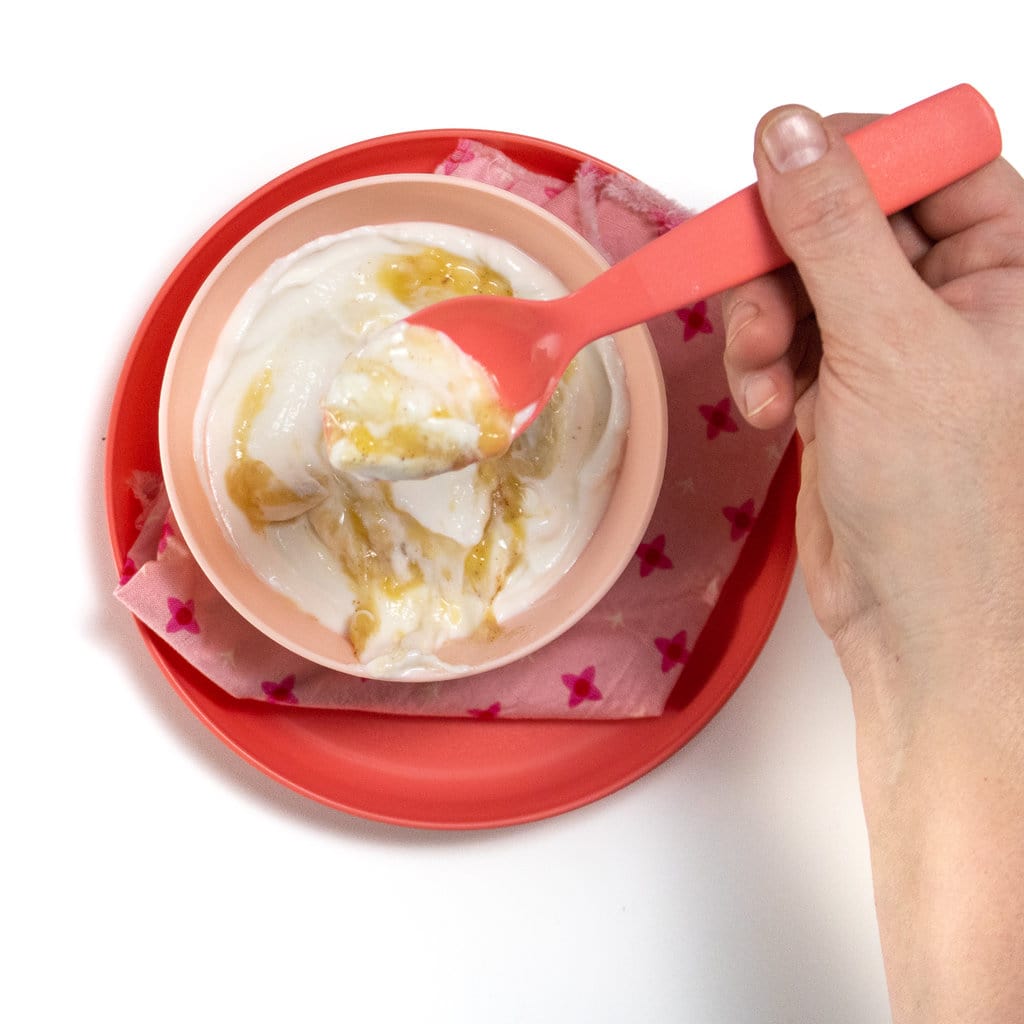 一只手拿着粉红色的汤匙在一个装满香蕉酸奶的粉红色儿童碗上。