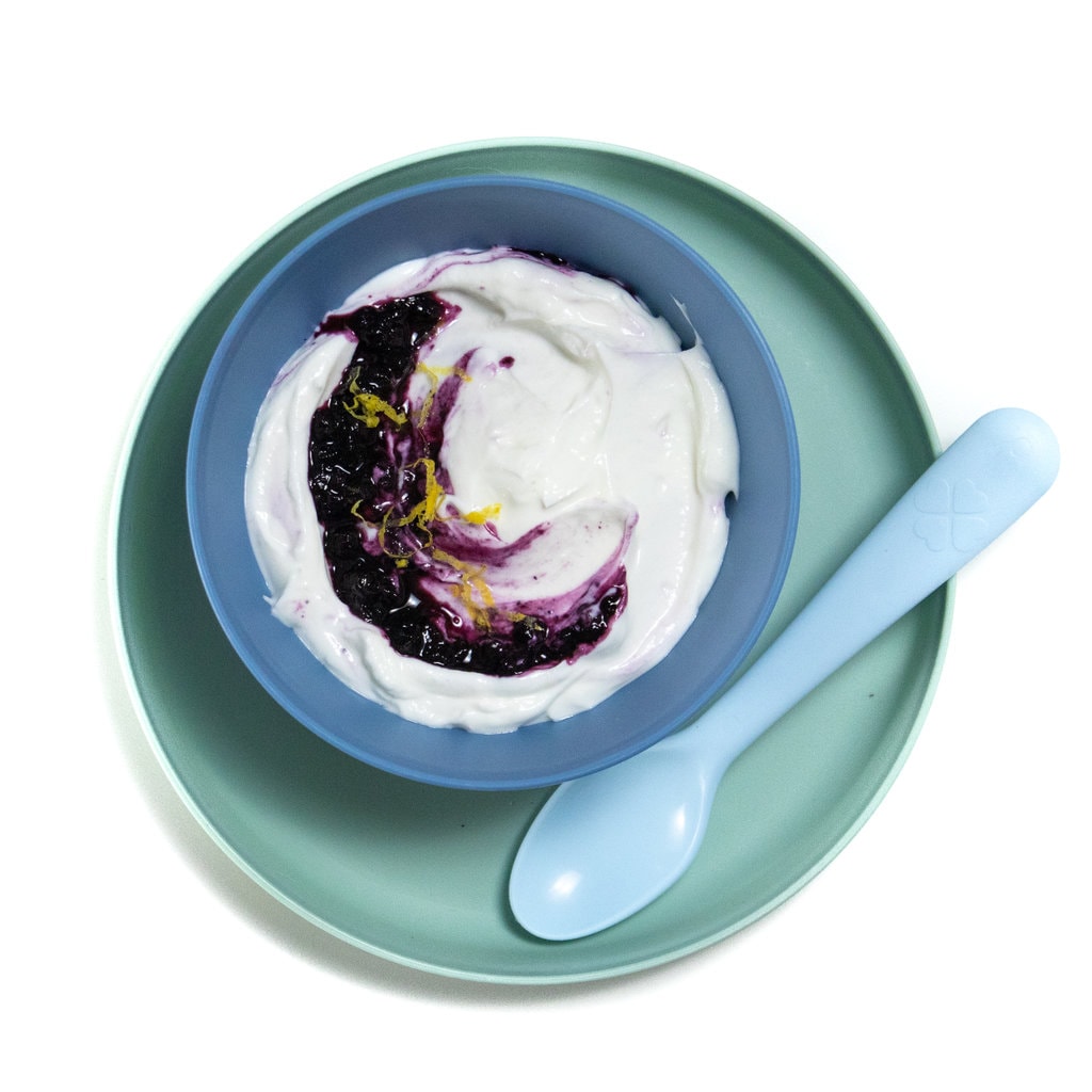 白色背景，带有蓝绿色的孩子盘，上面有一个蓝色的孩子碗，里面装满了蓝莓酸奶。