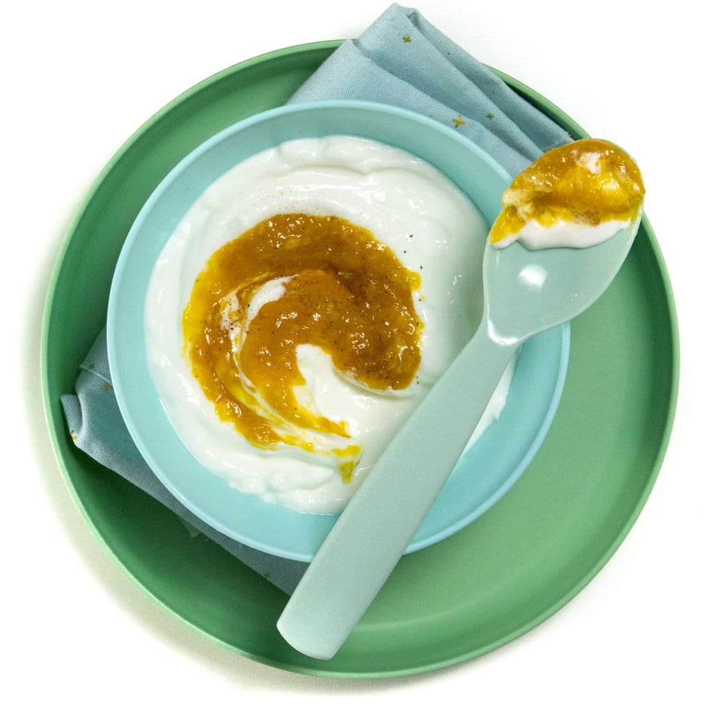 一个蓝色的儿童碗在绿色儿童板上，里面有芒果酸奶，勺子放在上面放着芒果酸奶。