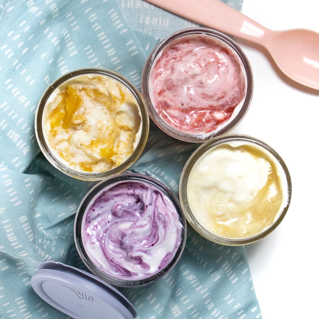 四个装满酸奶的玻璃罐，上面旋转着蓝色的餐巾，粉红色的勺子和蓝色的盖子在白色背景上。