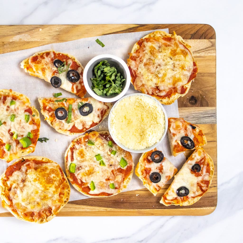 大理石台面上的木制切菜板，上面有几个不同的英式松饼披萨，带有不同的浇头，一个装满帕尔玛干酪和辣味罗勒的碗。