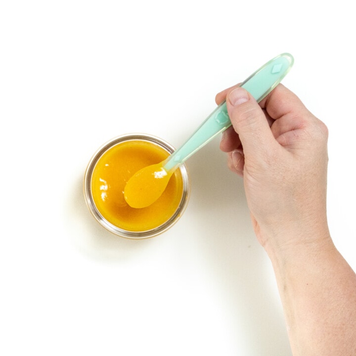 一只手拿着婴儿勺子在玻璃罐上拿着杏子食品泥，靠在白色背景上。