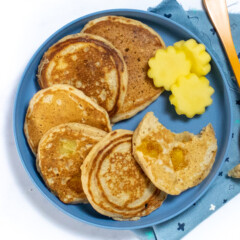 一个带有三个芒果煎饼和芒果的蓝色儿童盘子切成蓝色的彩色餐巾纸和白色背景的橙色叉子。
