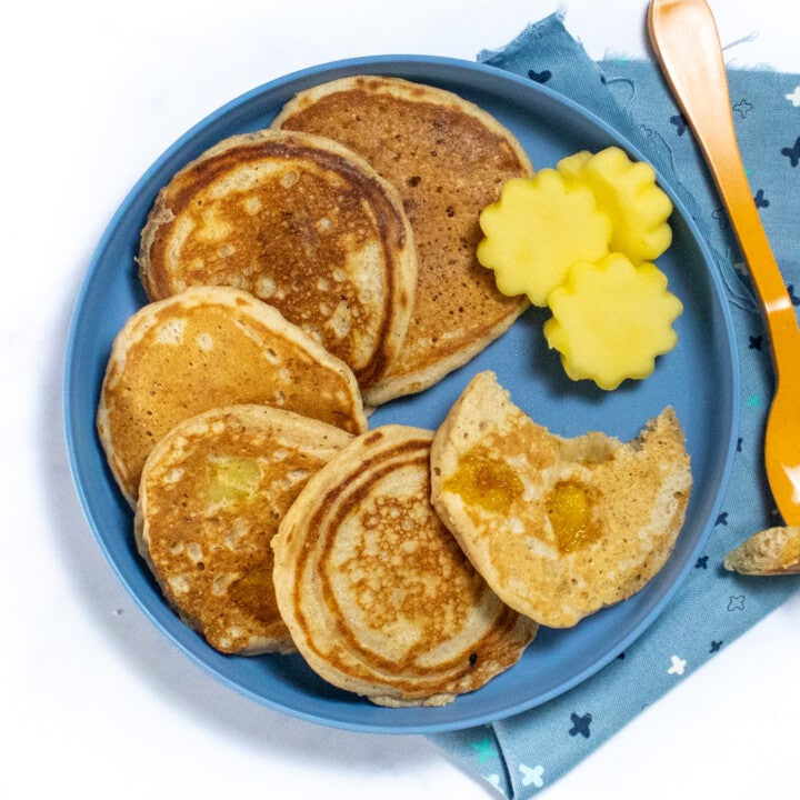 一个带有三个芒果煎饼和芒果的蓝色儿童盘子切成蓝色的彩色餐巾纸和白色背景的橙色叉子。