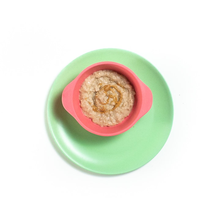 粉红色的婴儿碗在绿色盘子上。碗中是燕麦谷物和花生酱的漩涡。