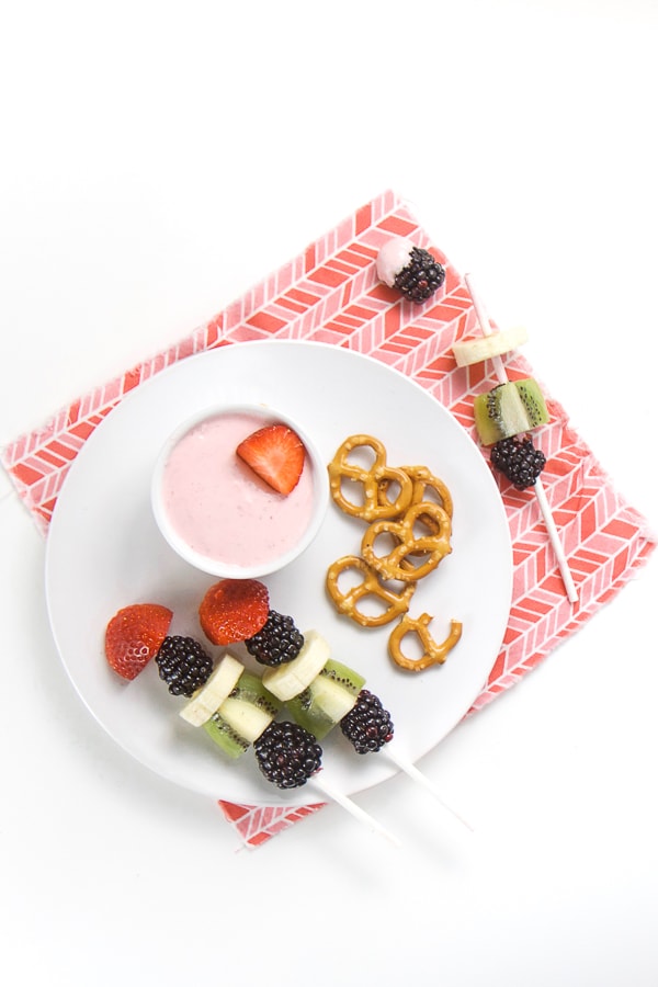 白板上装小碗草莓奶油芝士和2个果园加草莓、香蕉、kiwi树莓和黑莓以及一小堆retzel全上粉红色餐巾