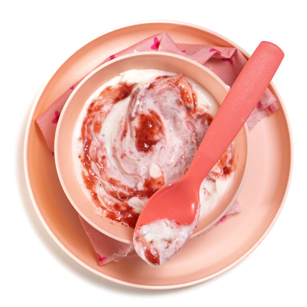 一个粉红色的儿童碗，上面有粉红色的餐巾纸和粉红色的勺子，拿着一个为婴儿，幼儿和孩子们的草莓酸奶。